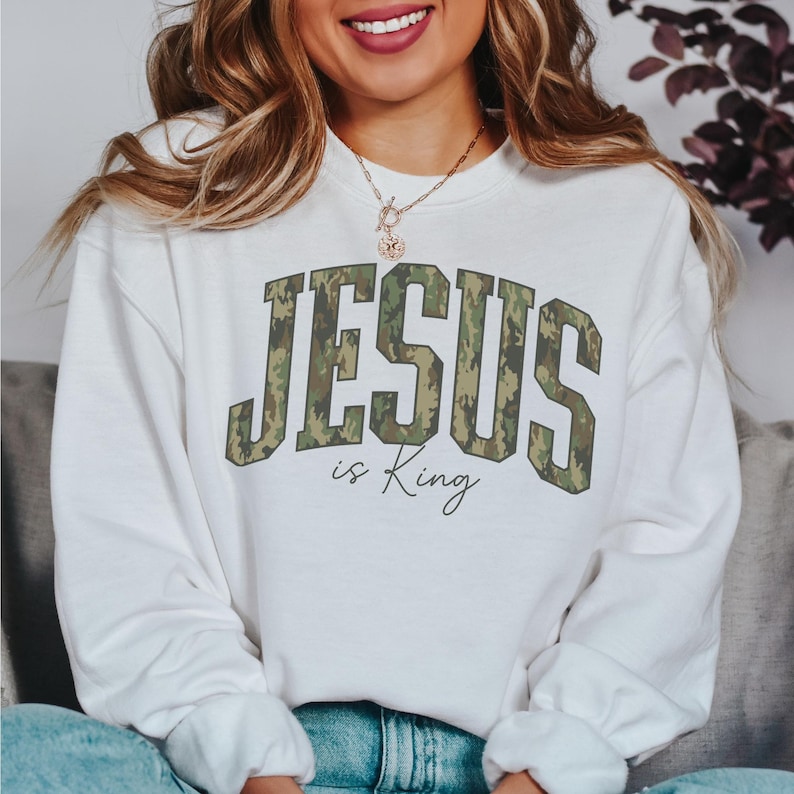 May include: White sweatshirt with the text "JESUS is King" in a camouflage design. The sweatshirt is a crew neck style and appears to be made of a soft material. The text is in a bold, collegiate-style font.