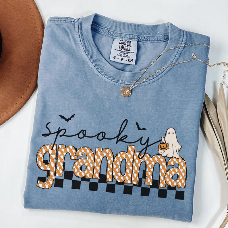 Op de afbeelding: Een lichtblauw T-shirt met de tekst "Spooky Grandma" in zwart en oranje. De tekst is omringd door een patroon van witte geesten met oranje pompoenen. Het shirt heeft ook twee zwarte vleermuizen en een witte geest die een pompoen vasthoudt.