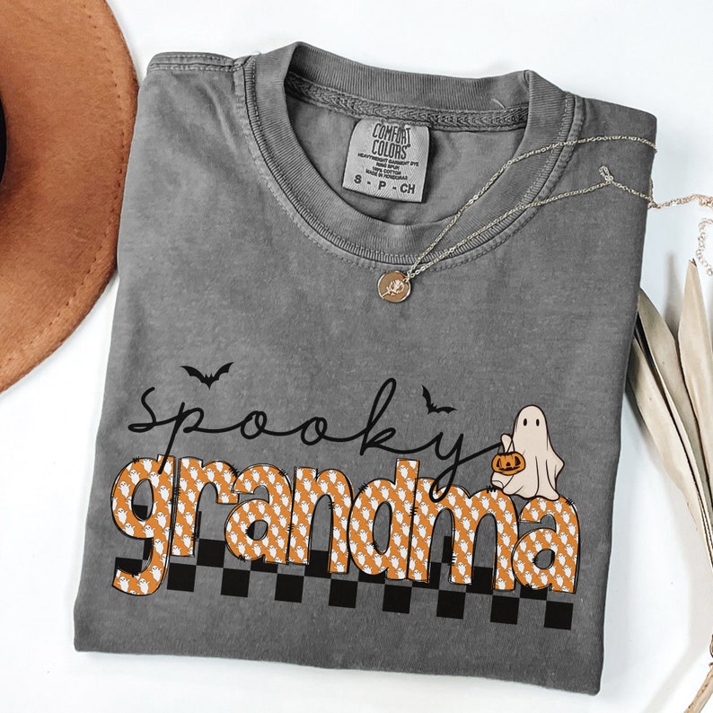 Op de afbeelding: Grijs T-shirt met een zwarte en oranje "Spooky Grandma" afbeelding. De afbeelding toont een geest die een pompoen vasthoudt en vleermuizen die boven de tekst vliegen.