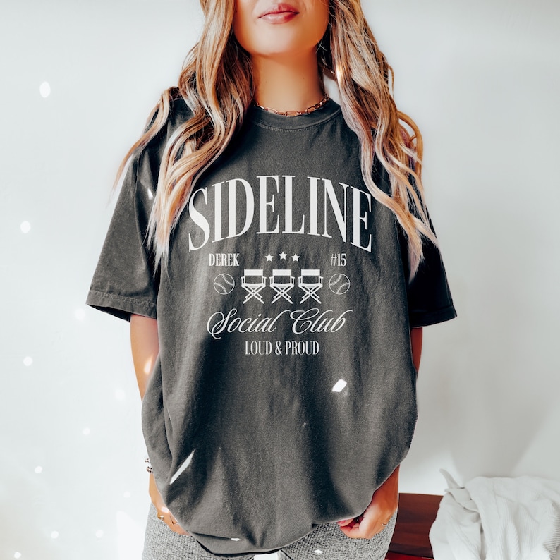 Puede incluir: Una camiseta gris oscuro con el texto "SIDELINE Social Club LOUD & PROUD" y un gr&aacute;fico de tem&aacute;tica de b&eacute;isbol. El gr&aacute;fico incluye una pelota de b&eacute;isbol, sillas de director y estrellas.