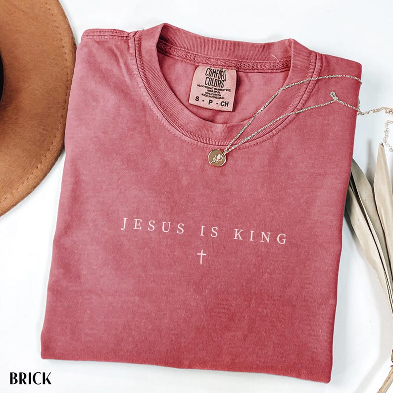 May include: Brick-colored Comfort Colors t-shirt with the text "JESUS IS KING" and a cross. A silver necklace with a round pendant rests on the shirt. The shirt is folded and placed on a white surface.