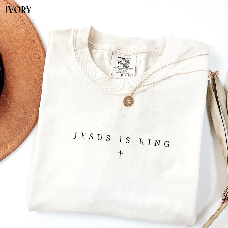 May include: Ivory-colored t-shirt with the text "JESUS IS KING" and a cross. A gold necklace with a round pendant rests on the shirt. The shirt has a Comfort Colors tag. A brown hat is partially visible.