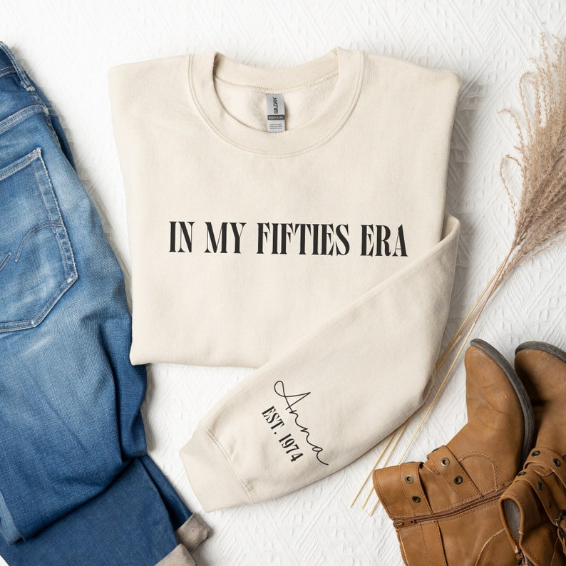 May include: A beige sweatshirt with the text "IN MY FIFTIES ERA" printed on the front and "Anna EST. 1974" printed on the sleeve.