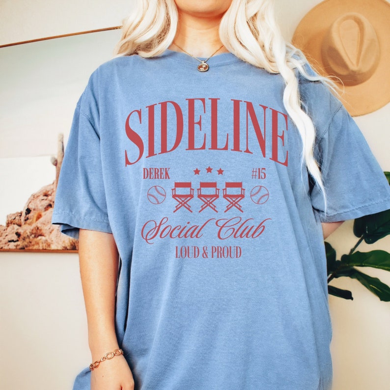 Puede incluir: Una camiseta azul claro con un gr&aacute;fico rojo que dice "Sideline Social Club" con el texto "Derek #15" y cuatro sillas de director rojas. El texto "Loud & Proud" est&aacute; debajo del gr&aacute;fico.