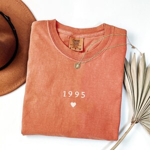 May include: A terracotta-colored t-shirt with the year 1995 and a small white heart printed on the front. The shirt is made by Comfort Colors.