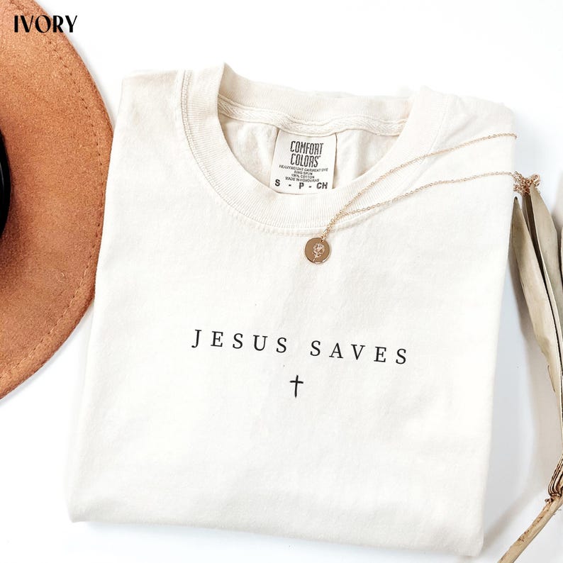 Puede incluir: Camiseta de color marfil con el texto "JESUS SAVES" y una cruz. Un collar dorado con un colgante redondo descansa sobre la camiseta. La camiseta tiene una etiqueta Comfort Colors. Un sombrero marr&oacute;n y hojas secas est&aacute;n en el fondo.