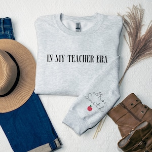 Pu&ograve; includere: Felpa grigia con il testo "IN MY TEACHER ERA" sul davanti e "Mrs. Smith" con una mela rossa sulla manica.