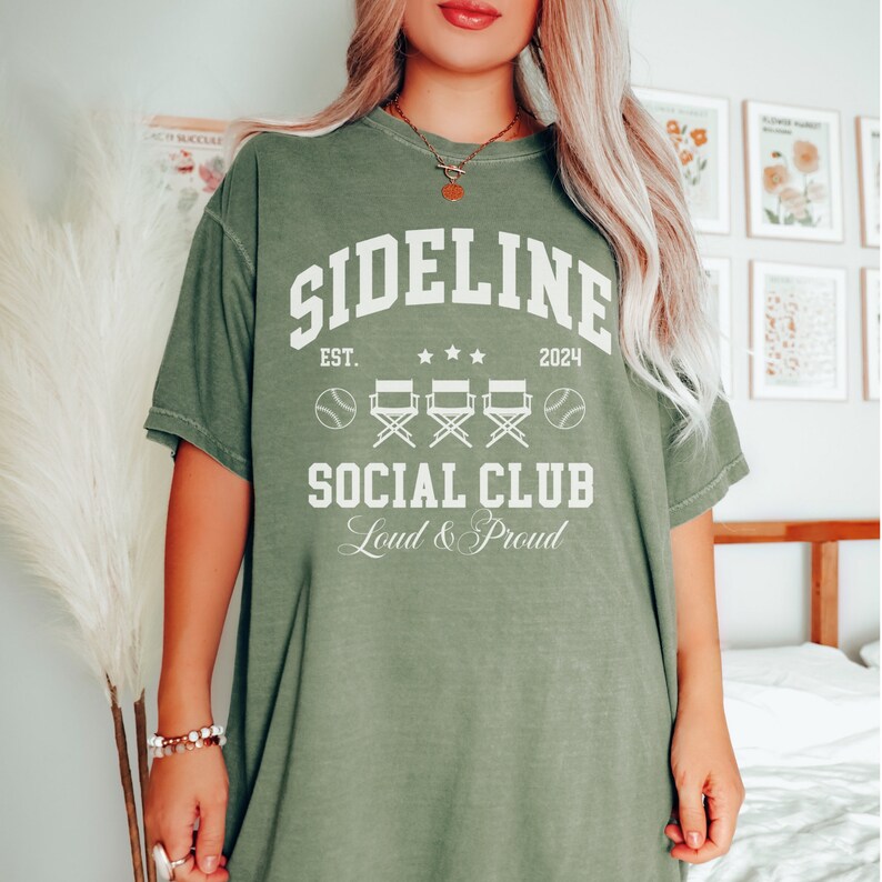 Op de afbeelding: Een groen T-shirt met witte tekst die "Sideline Social Club Est. 2024 Loud & Proud" luidt. De tekst wordt omringd door honkballen en regisseursstoelen.
