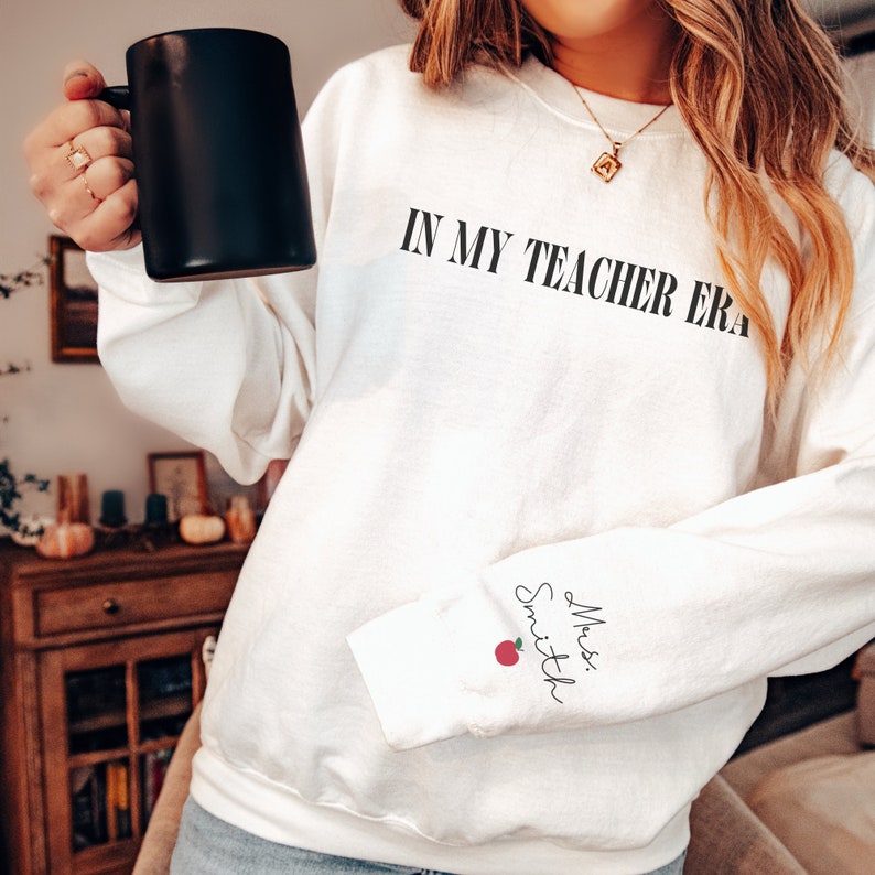 Pu&ograve; includere: Felpa bianca con la scritta "In My Teacher Era" stampata sul davanti e "Mrs. Smith" con una mela rossa stampata sulla manica sinistra.