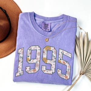 May include: A purple t-shirt with a floral design in the numbers "1995". The shirt is folded and laid on a white surface.