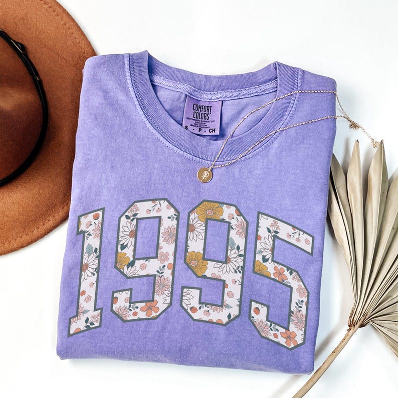 May include: A purple t-shirt with a floral design in the numbers "1995". The shirt is folded and laid on a white surface.