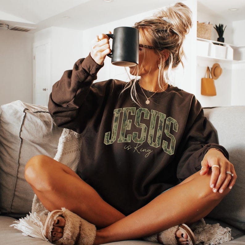 May include: A brown sweatshirt with the text "JESUS is King" in a camouflage design. The person is holding a black mug. The sweatshirt is long-sleeved and the person is wearing fuzzy slippers.