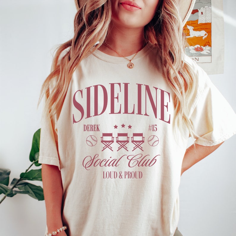 Puede incluir: Una camiseta de color crema con un gr&aacute;fico rosa que dice "Sideline Social Club" con el texto "Derek #15" y tres sillas de director con pelotas de b&eacute;isbol a cada lado. El texto "Loud & Proud" est&aacute; debajo del gr&aacute;fico.