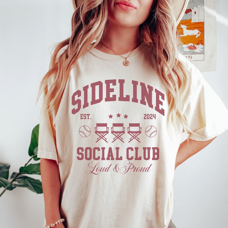 Op de afbeelding: Een cr&egrave;mekleurig T-shirt met een kastanjebruine afbeelding met de tekst "Sideline Social Club" met de tekst "Est. 2024" en drie regisseursstoelen met honkballen aan beide kanten.