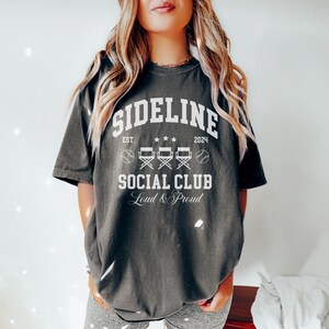 Op de afbeelding: Een donkergrijs T-shirt met een witte afbeelding waarop "Sideline Social Club" staat, met daaronder de tekst "Est. 2024" en "Loud & Proud". De afbeelding bevat ook twee honkbalballen en vier regisseursstoelen.
