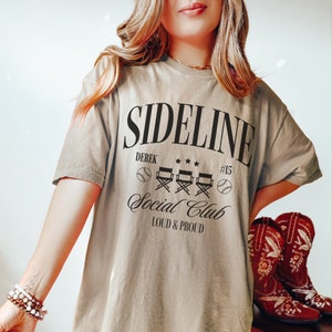 Puede incluir: Una camiseta beige con el texto "Sideline Social Club Loud & Proud" y un gr&aacute;fico de b&eacute;isbol. El texto "Derek #15" tambi&eacute;n est&aacute; en la camiseta.