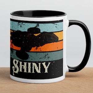 May include: White ceramic mug with a black interior and handle. The mug features a retro design with a black silhouette, orange and blue stripes, and the word "SHINY" in a stylized font.