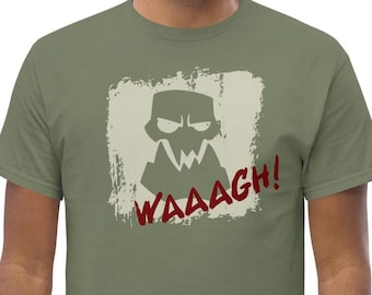Puede incluir: Camiseta verde oliva con un gráfico desgastado de un monstruo estilizado y la palabra "Waaagh!" en rojo. El diseño está dentro de un marco rectangular blanquecino descolorido. La camiseta es de material suave.