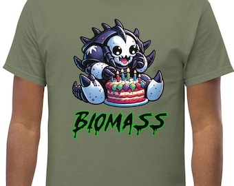 Puede incluir: Una camiseta de color verde oliva con el gráfico de una criatura de dibujos animados con armadura oscura y detalles blancos, sentada frente a un pastel de cumpleaños colorido con velas encendidas. Debajo, la palabra BIOMASS en texto verde.