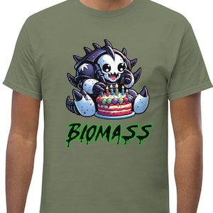 Puede incluir: Una camiseta verde con una ilustración de dibujos animados de una criatura en blanco y negro comiendo un pastel de cumpleaños con velas. El texto "BIOMASS" está escrito en verde debajo de la criatura.