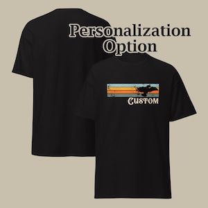 Op de afbeelding: Een zwart T-shirt met korte mouwen voorzien van een afbeelding met horizontale strepen in blauw, oranje en beige, een zwart silhouet van een voertuig en het woord CUSTOM eronder gedrukt. De tekst Personalization Option is boven het T-shirt zichtbaar.