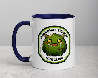 Puede incluir: Taza de café de cerámica blanca con borde azul. La taza presenta un monstruo de dibujos animados verde con ojos amarillos y una boca que gotea. El monstruo está dentro de un octógono blanco con el texto "Emotional Support Nurgling" en negro.