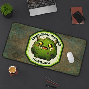 May include: A rectangular desk mat with a distressed green and brown pattern. The mat features a cartoon monster with the text "EMOTIONAL SUPPORT NURGLING" in a green and white badge. A laptop, mouse, and other office supplies are visible.