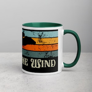 May include: White ceramic mug with a green rim and handle. The mug features a retro-style design with orange, yellow, and blue stripes and a silhouette of a person running. The text "Ride the Wind" is printed in black at the bottom of the mug.
