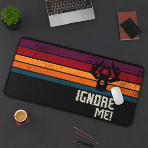 Könnte beinhalten: Schwarze rechteckige Schreibtischunterlage mit Retro-Design. Die Matte hat horizontale Streifen in Lila, Rot, Orange, Blau und Schwarz. Eine schwarze Silhouette und die Worte "IGNORE ME!" sind auf der Matte aufgedruckt.