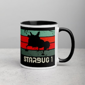 Op de afbeelding: Witte keramische mok met een zwarte rand en handvat. De mok heeft een retro-ontwerp met rode, groene en witte strepen. Een zwart silhouet van een ruimteschip bevindt zich in het midden van de strepen. De tekst "STARBUG 1" is in wit onder het ruimteschip gedrukt.