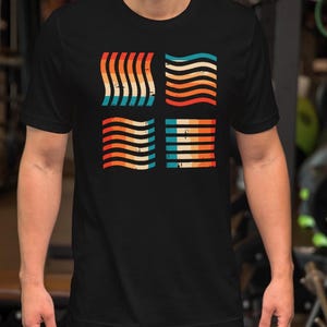 May include: Black t-shirt with a retro design featuring four squares. Each square has wavy horizontal lines in orange, red, blue, and cream. The shirt is made of a soft material.
