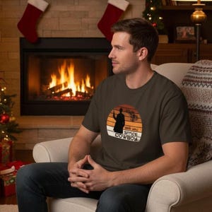 May include: A dark brown t-shirt featuring a silhouette of a figure holding a weapon against a sunset backdrop. The text "SER YOU SPACE COWBOY" is printed below the image. The shirt is worn by a person sitting in a chair.