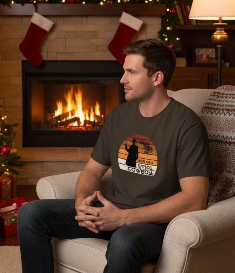 May include: A dark brown t-shirt featuring a silhouette of a figure holding a weapon against a sunset backdrop. The text "SER YOU SPACE COWBOY" is printed below the image. The shirt is worn by a person sitting in a chair.