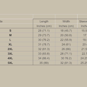 Op de afbeelding: Een beige maattabel voor kleding met lengtes, breedtes en mouwlengtes in inches en centimeters voor de maten S tot 5XL. De tabel bevat kolommen voor lengte, breedte en mouwlengte met specifieke numerieke waarden voor elk kledingstuk.