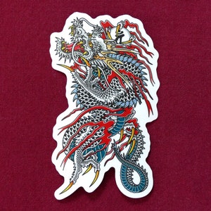 May include: A white and blue dragon tattoo sticker with red and black accents. The dragon is curled up with its head facing forward and its wings spread out. The sticker is on a red background.