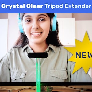May include: A green and black tripod extender with a clear plastic tube and a black base. The extender is on a table in front of a television screen showing a woman wearing headphones. The screen also has a yellow star with the word "NEW!" on it.