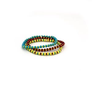 May include: Three circular beaded bracelets stacked together. One bracelet features bright turquoise beads, another consists of deep maroon beads, and the third is made of vibrant yellow beads. Each bracelet includes a single small metallic bead.