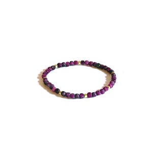 May include: A circular bracelet composed of small, faceted, deep magenta-colored beads interspersed with tiny, smooth, metallic gold-toned cube spacers. The beads have a textured, matte appearance, and the bracelet is arranged in a simple, continuous loop.