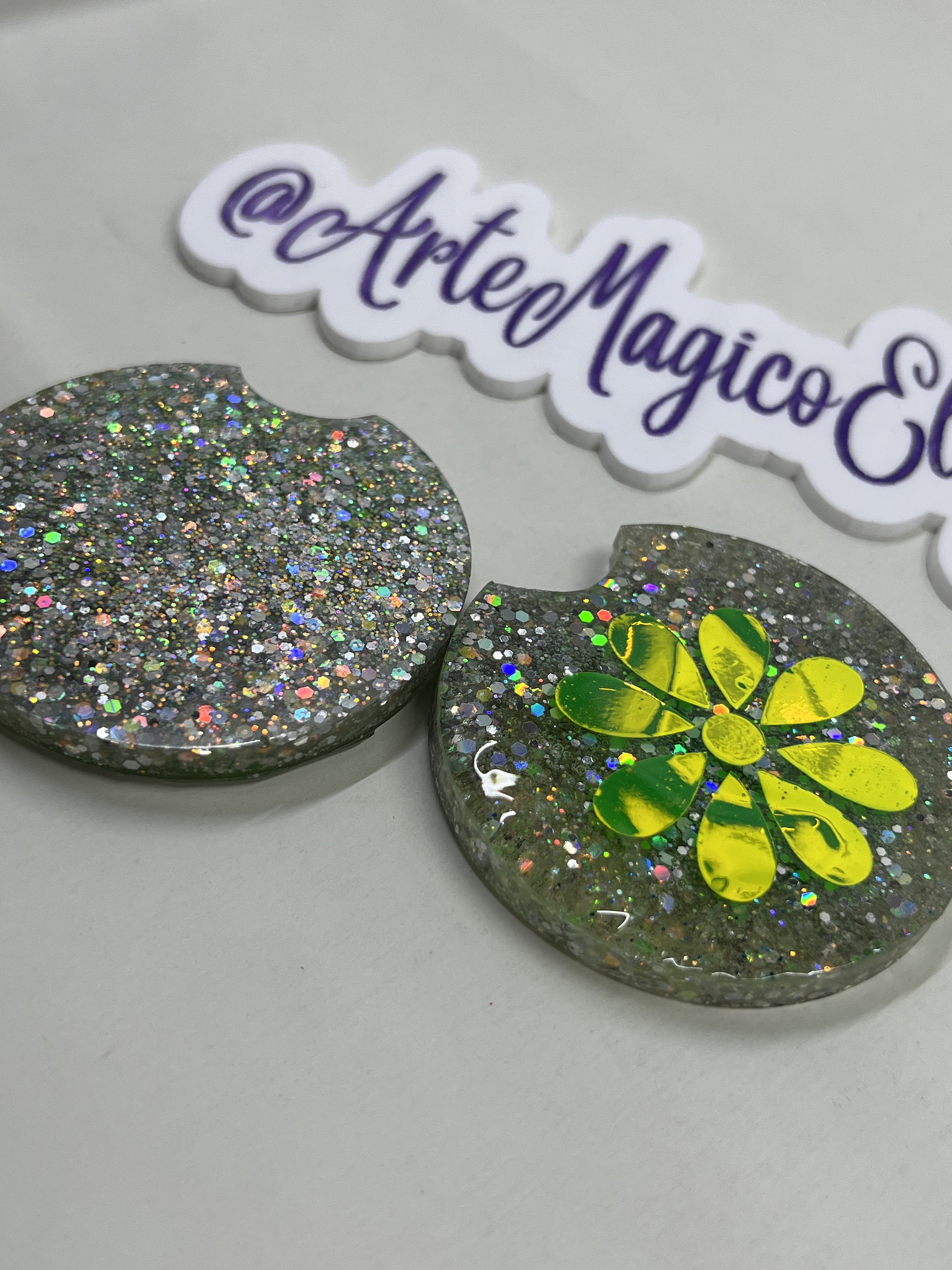 May include: Two round, silver glitter coasters. One coaster has a green and yellow flower design.