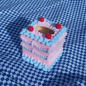 May include: A pink and blue cake-shaped tissue box holder with a wooden top and four red cherries on top.
