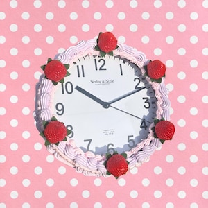 May include: A round wall clock with a white face, black numerals, and black hands. The frame is decorated with light pink piped frosting and five red strawberry embellishments. The text Sterling & Noble Clock Company is printed on the face.
