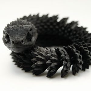 Puede incluir: Una figura de serpiente articulada en color negro mate enrollada en forma circular. La serpiente presenta una cabeza detallada con grandes ojos oscuros y un cuerpo cubierto de numerosas protuberancias puntiagudas similares a escamas a lo largo de toda su longitud.