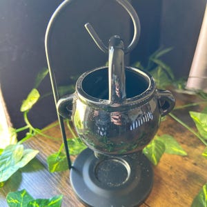 May include: A black ceramic cauldron-shaped incense holder with a handle and two side handles. The incense holder sits on a black metal stand with a curved arm. Green leaves surround the incense holder.