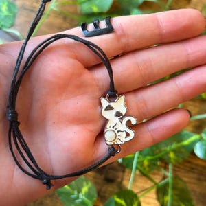 May include: A black cord bracelet featuring a white cat charm with black spots and a sun design. The cat charm is made of metal and has a glossy finish. The bracelet is adjustable.