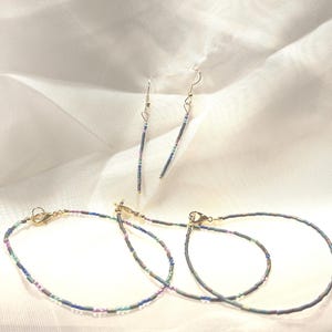 May include: A set of jewelry consisting of two delicate bracelets and a pair of matching drop earrings. Each piece is crafted from small, iridescent seed beads in shades of blue, purple, and green, finished with gold-toned metal clasps and hooks.
