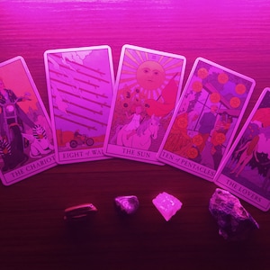 May include: Five tarot cards laid out on a wooden surface under a pink light. The cards are "The Chariot", "Eight of Wands", "The Sun", "Ten of Pentacles", and "The Lovers".