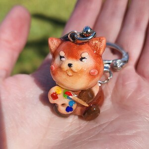 May include: A small, brown and white ceramic keychain shaped like a dog holding a paint palette with colorful paint. The keychain has a silver ring.