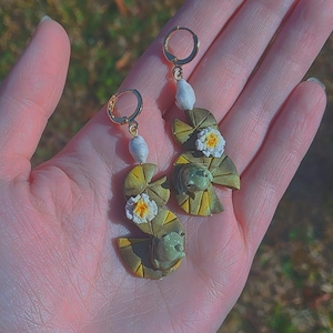 May include: A pair of gold-colored hoop earrings featuring lily pad and frog designs. Each earring has a white pearl-like bead and a lily pad with a small white and yellow flower. The earrings are held in a hand.