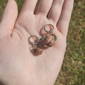 Puede incluir: Un par de pendientes de aro dorados con colgantes de tetera de arcilla marrón. Las teteras tienen hojas verdes y cuelgan de los aros.