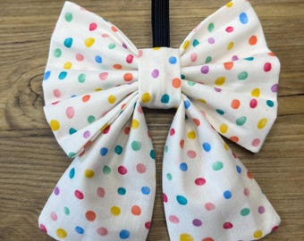 May include: A white fabric bow tie with a colorful polka dot pattern. The dots are in shades of red, orange, yellow, green, blue, and purple. The bow tie has a black elastic band for attachment. The bow tie is likely for a pet or child.